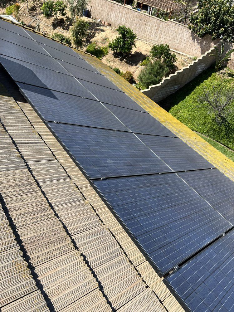 A high-angle view of black solar panels installed on a residential tile roof, with a grassy yard and stone wall below.