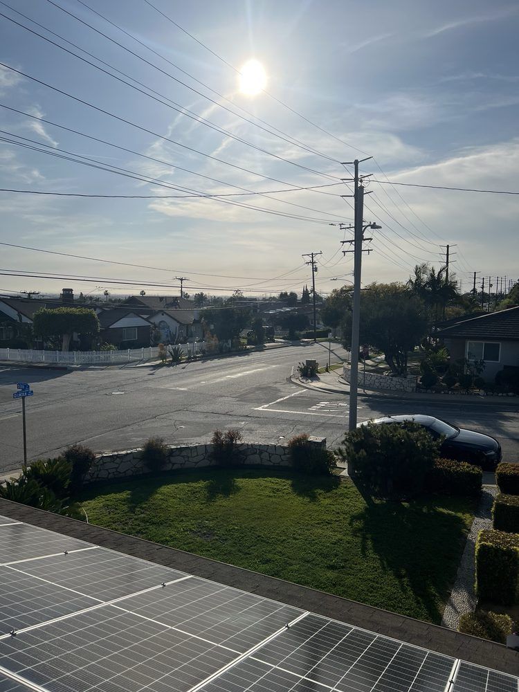 A bright sun shines over a suburban street, a grassy lawn, and solar panels on a roof in the foreground.