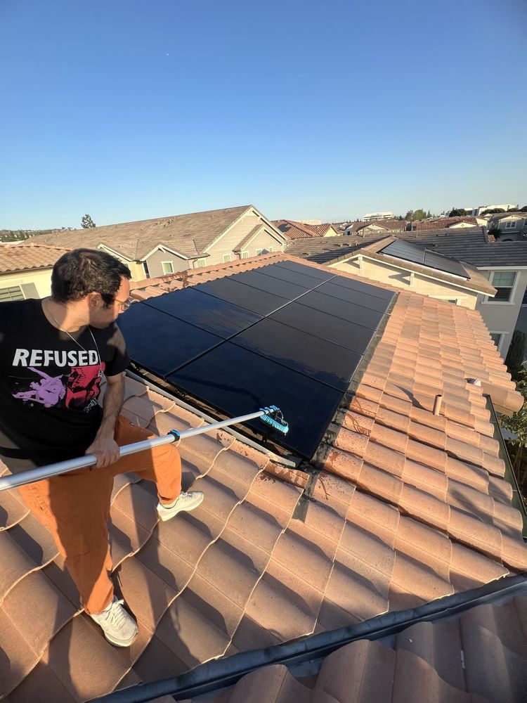 A person in an orange shirt uses a long brush to clean solar panels on a brown-tiled roof under a clear blue sky.