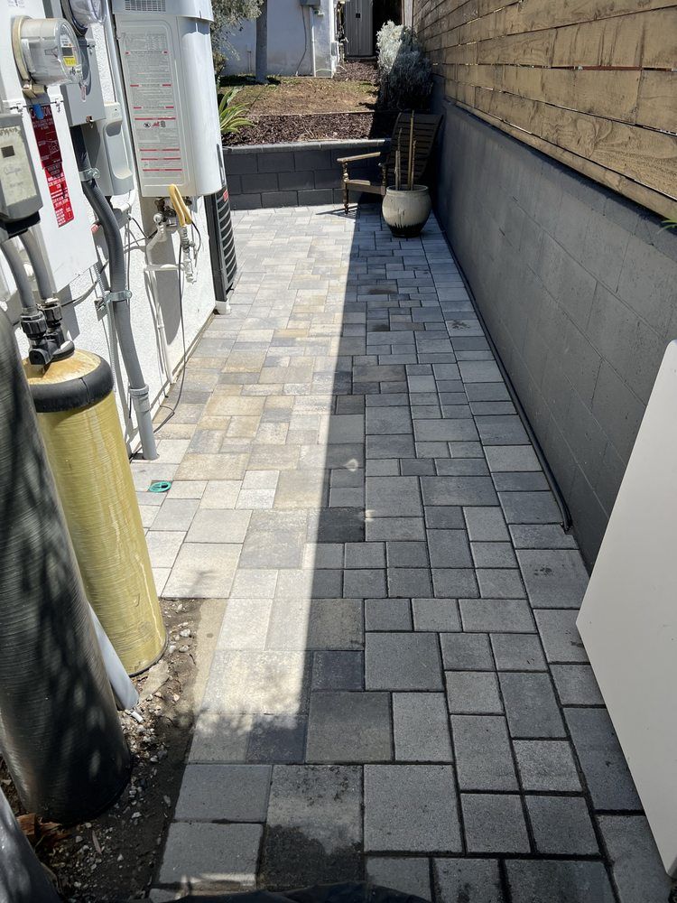 A side path featuring light and dark gray stone pavers, bordered by a concrete wall on the right and utility equipment.