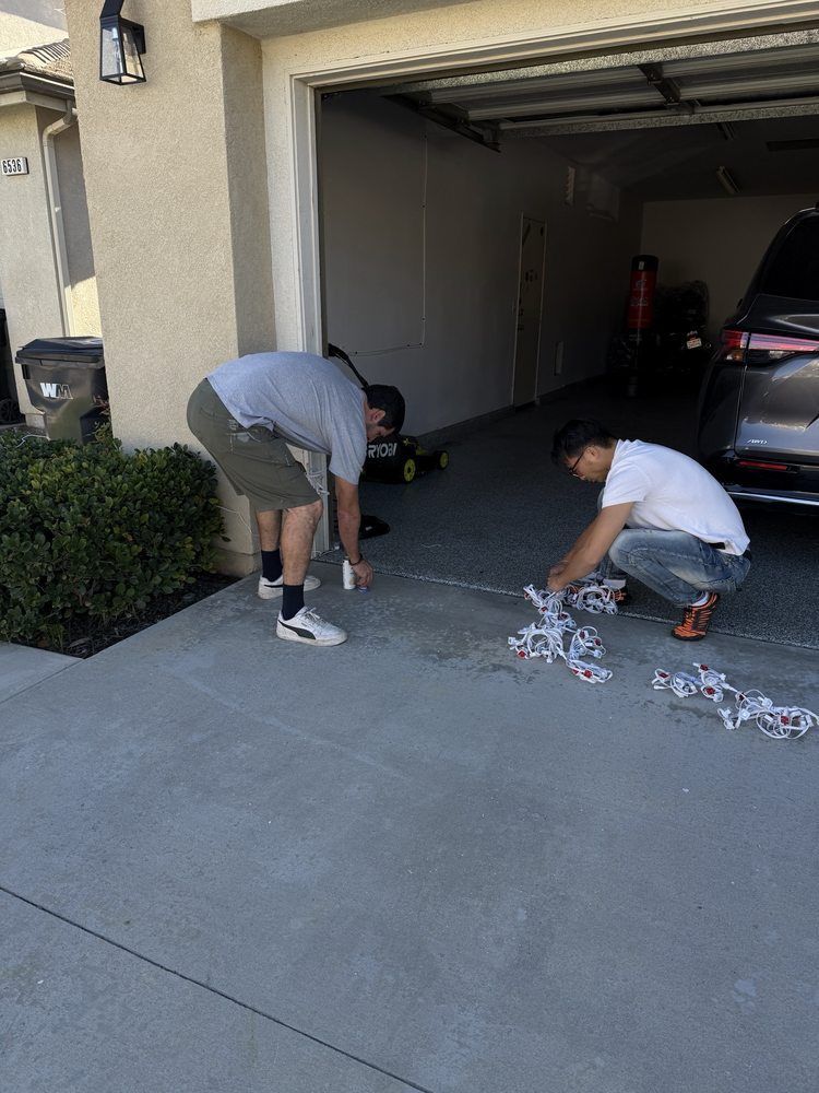 Two people kneeling on a driveway, applying tape to the concrete floor near the garage opening.