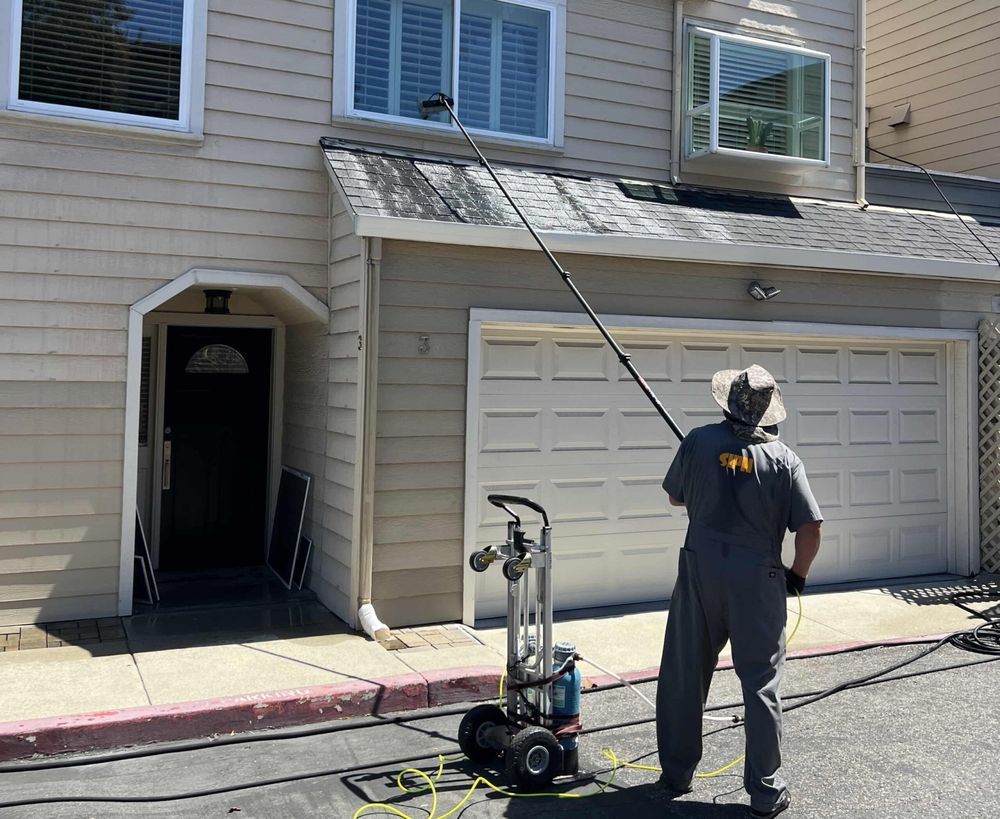 A person in uniform uses a long pole tool to wash the second-story windows of a beige house.