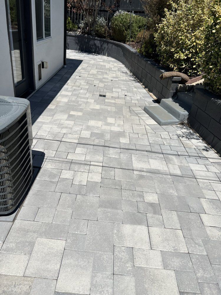 A grey, multi-sized paver stone walkway runs alongside a house exterior, leading toward a dark stone retaining wall.