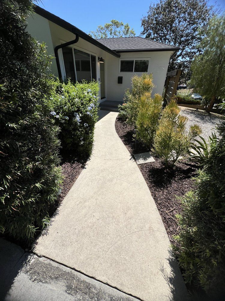 A concrete walkway leads through a landscaped garden toward the entrance of a one-story, cream-colored house.