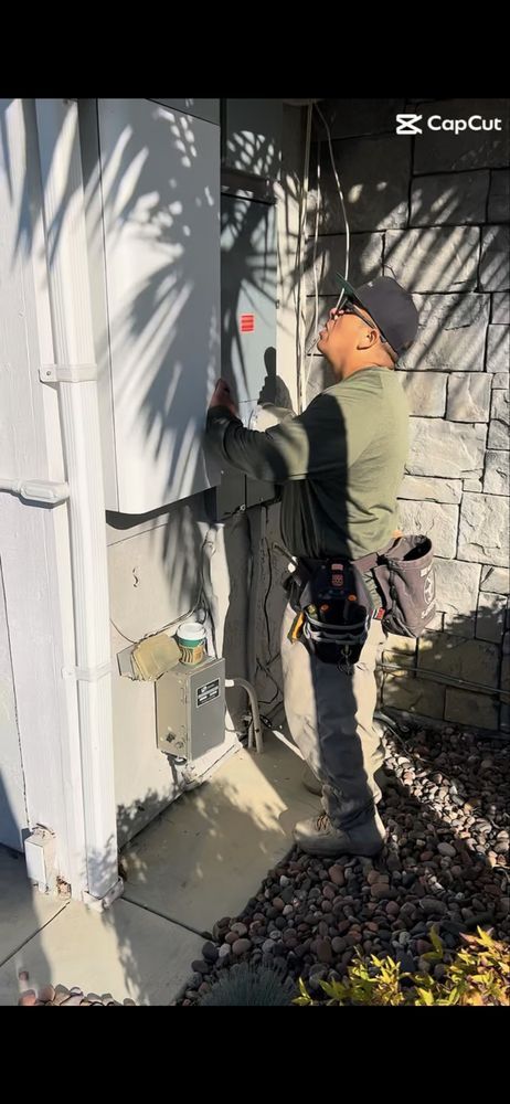 A person in a work vest and tool belt inspects an electrical panel mounted on a stone-clad outdoor wall.