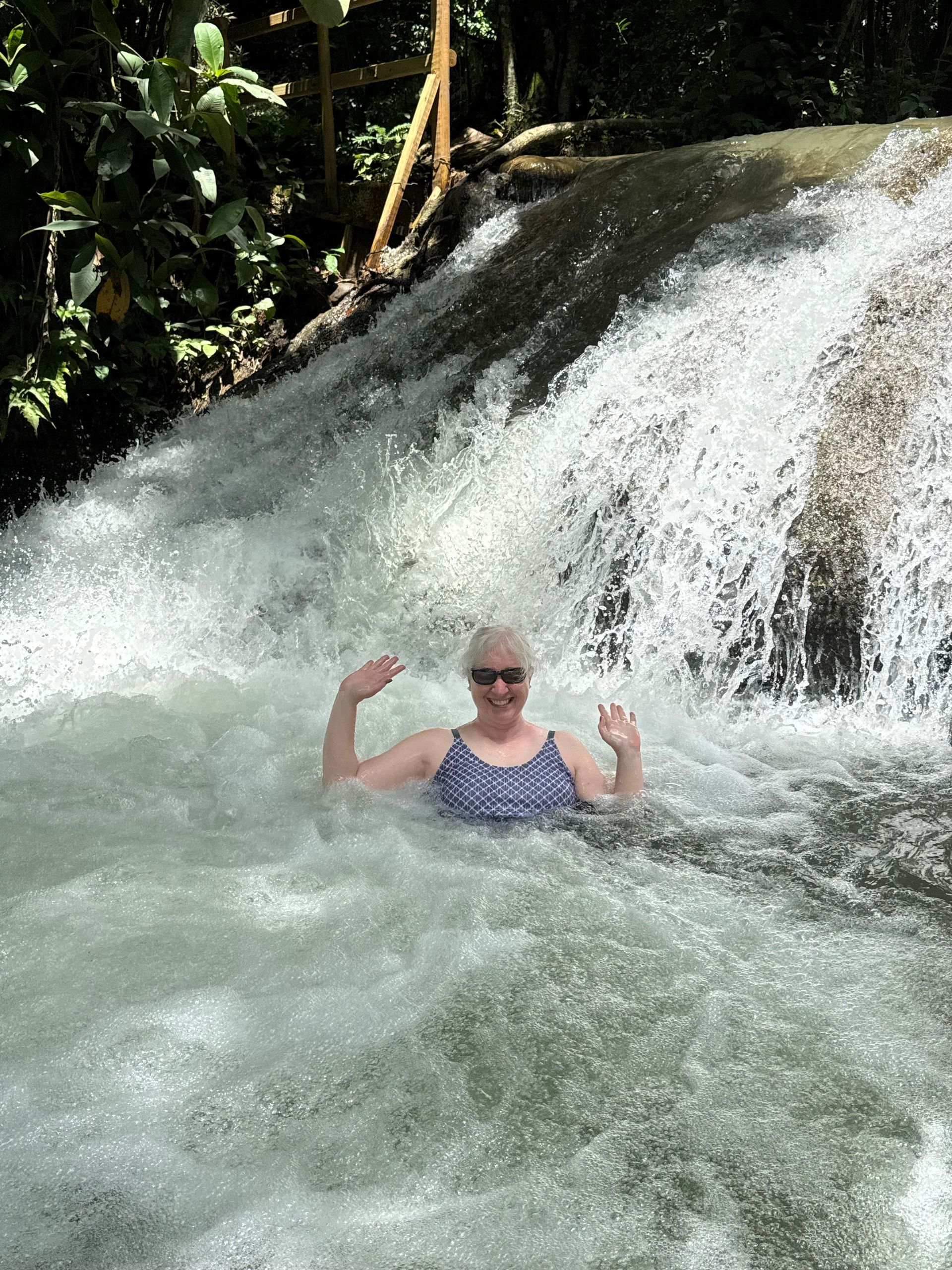 Woman in blue swimsuit waves from rushing waterfall.