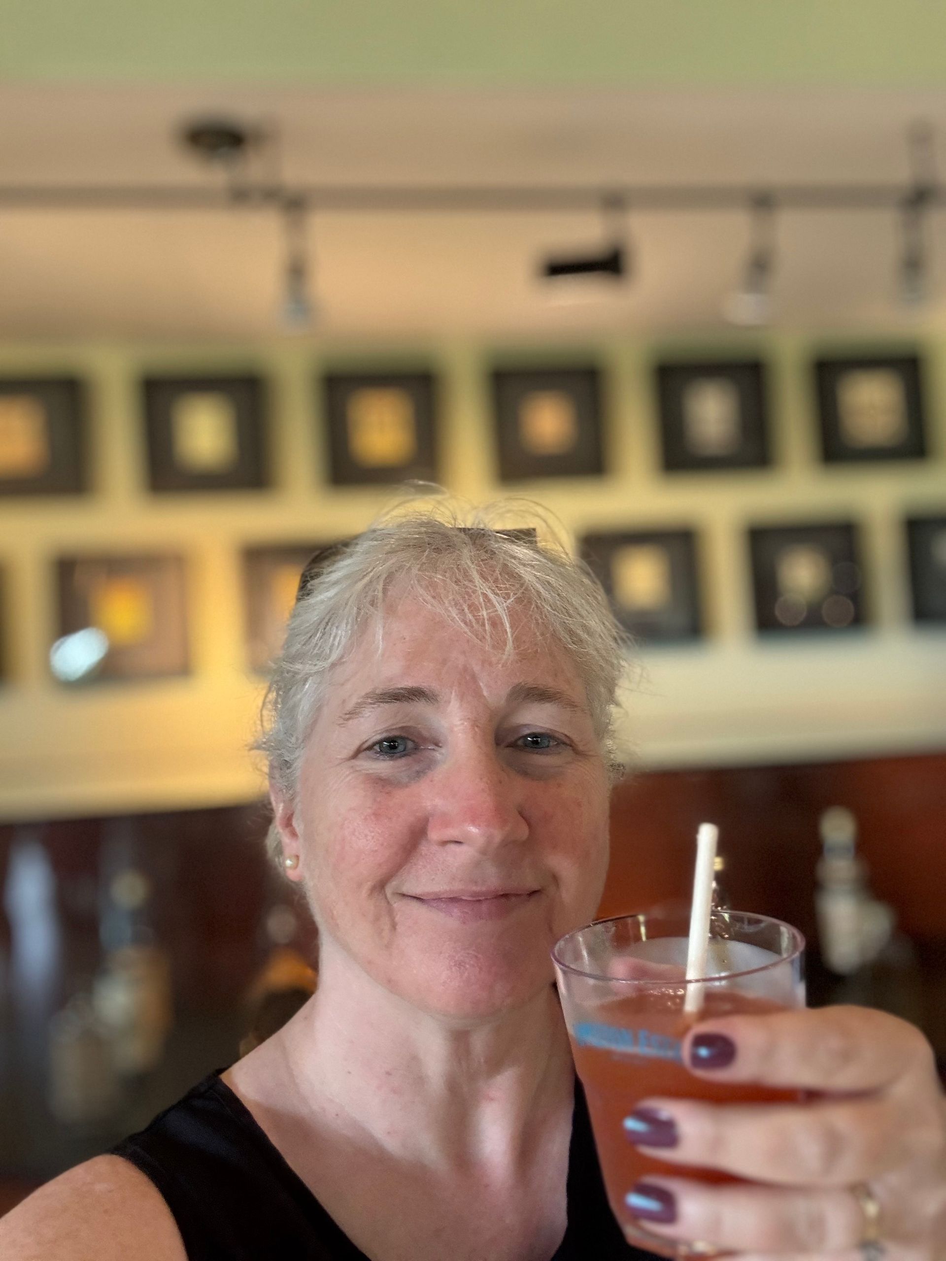 Woman holding a pink drink, smiling. In a room with framed art and a bar.