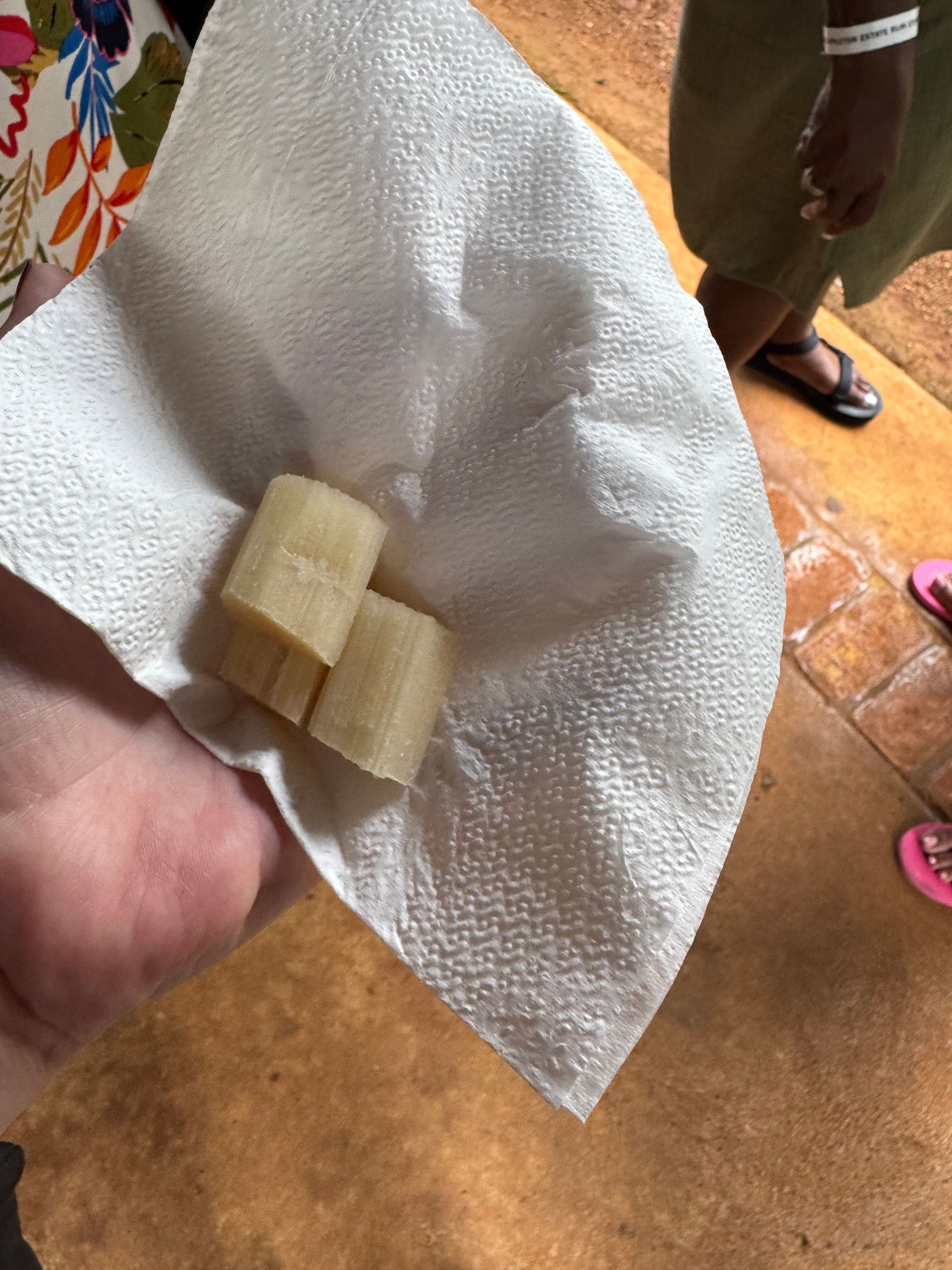 Hand holding a napkin with two pieces of light-colored sugar cane. Tan floor and feet wearing sandals in the background.