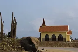 A small yellow church with a red roof and a large rock in front of it.