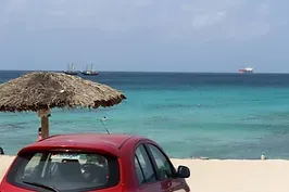 A red car is parked on the beach next to a thatched umbrella.