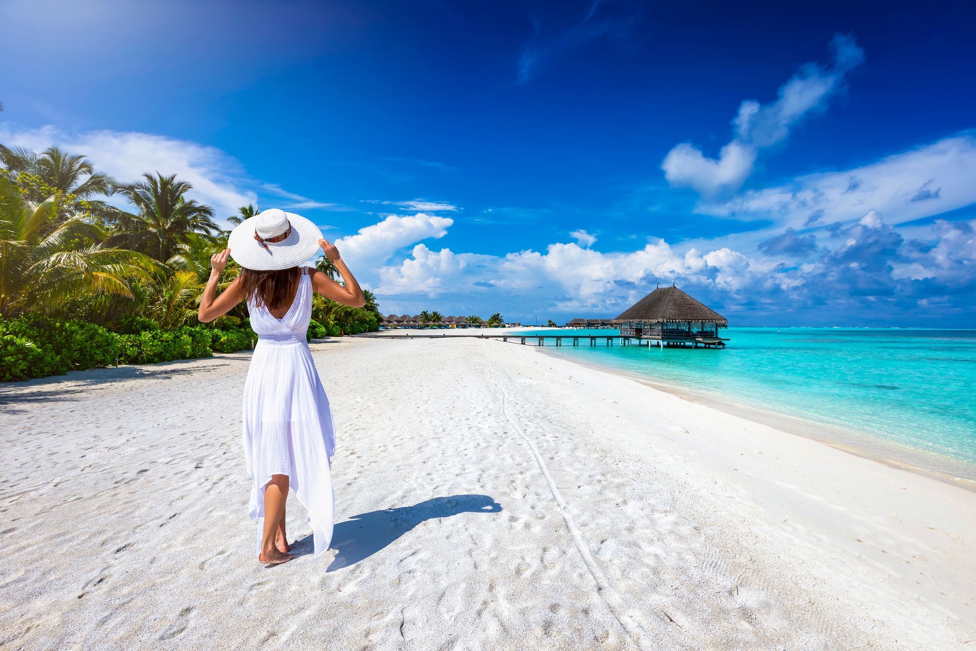 A woman in a white dress and hat is walking on a tropical beach.
