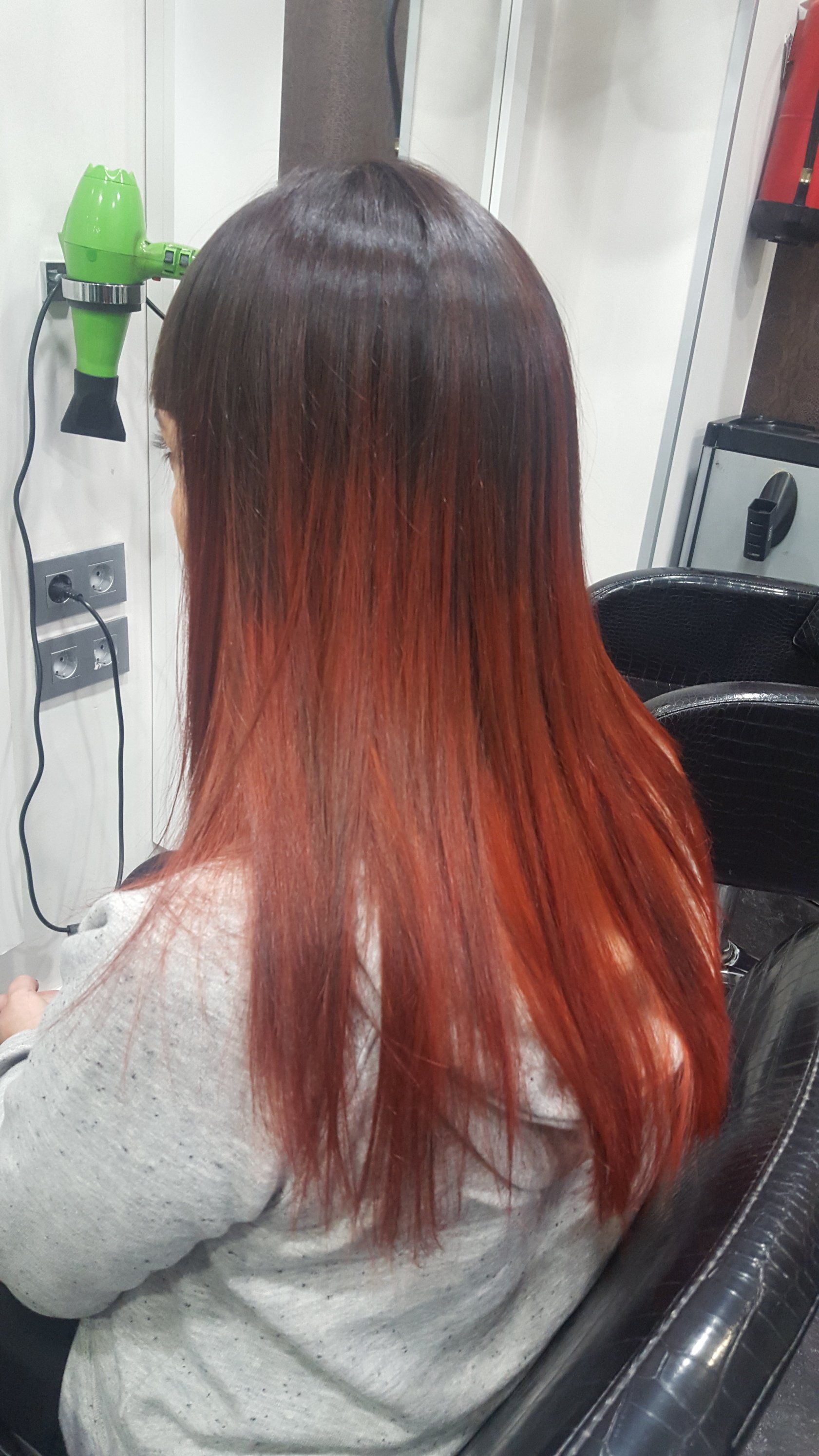 Una mujer con cabello largo y rojo está sentada en una silla frente a un secador de pelo verde.