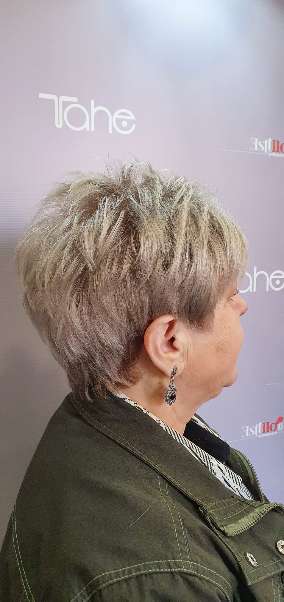 Una mujer con cabello rubio corto lleva una chaqueta verde y pendientes.