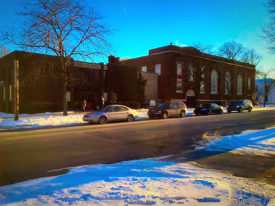 Cars are parked on the side of the road in front of a building covered in snow.