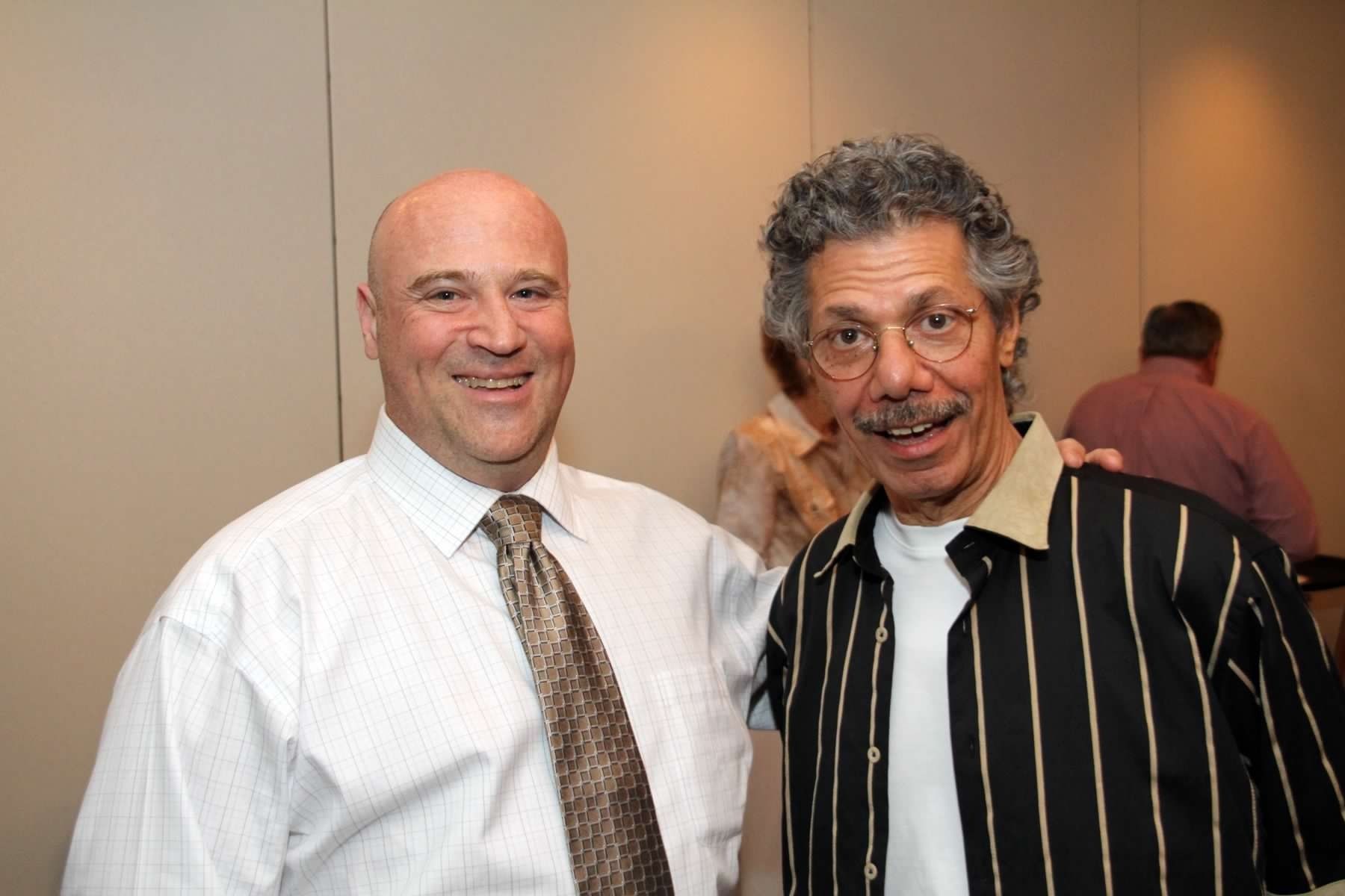 Two men smiling, one bald in a white shirt and tie, the other with curly gray hair, a striped shirt, and glasses.