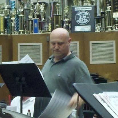 Man in green shirt reading music in front of trophies, likely in a band room.