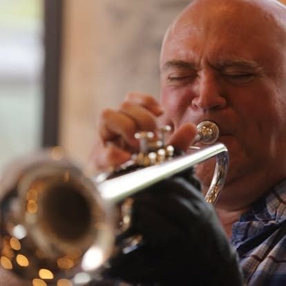 Bald man playing a silver trumpet, eyes closed, indoor setting.
