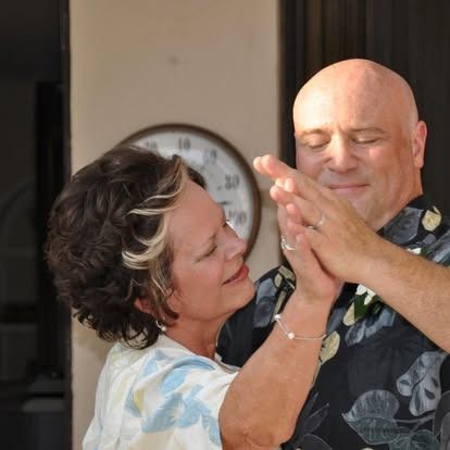 Couple dancing, man bald in Hawaiian shirt, woman with short hair, clock on wall.
