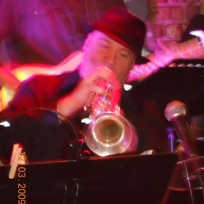 Man playing trumpet onstage, wearing a fedora. Blurred red and purple stage lights.