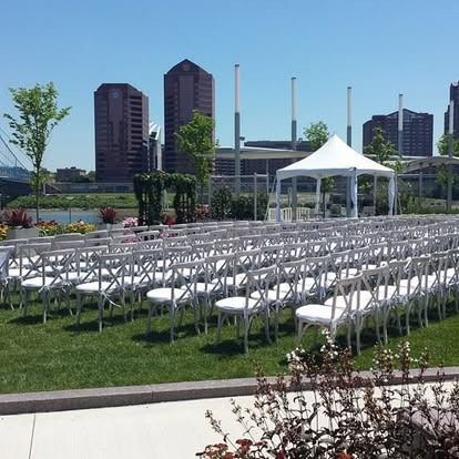 Rows of white chairs set up for a wedding ceremony in front of a tent, with city buildings in the background.