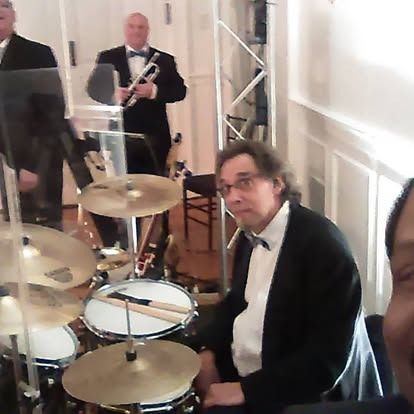 Band playing music at an event. Drummer, trumpet player, and another musician in a room.