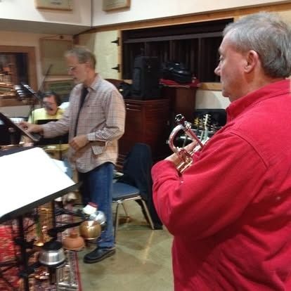Three men in a recording studio. One plays a trumpet, one reads music, and one is in the background.