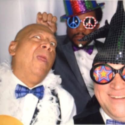Three men in silly outfits, photo booth setting. One makes a face, another wears peace sign glasses, all smiling.