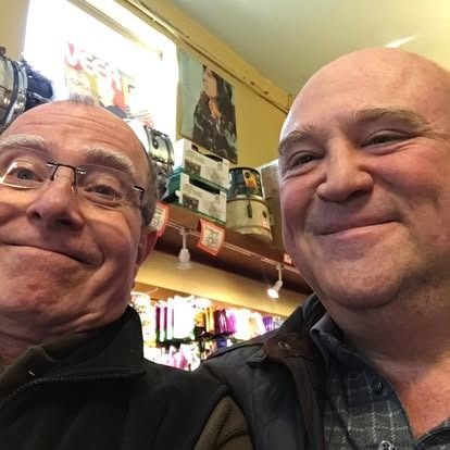 Two men smile at the camera, inside a store with shelves and products in the background.