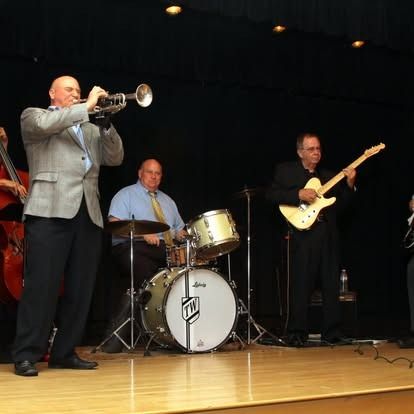 Jazz band on stage: trumpet player, drummer, guitarist, bassist; black backdrop.