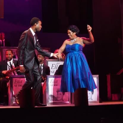 A man and woman dance on stage, she wears a blue dress, he a suit, with band in the background.