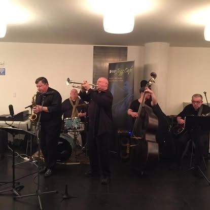Jazz band performing indoors: saxophone, trumpet, drums, upright bass, and guitar, all in black attire.