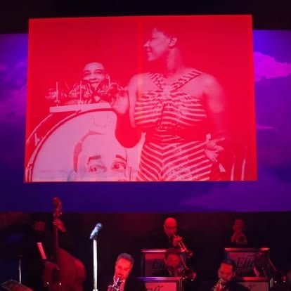 Stage performance: jazz band in front of a red-tinted screen displaying vintage imagery of a woman and a man.