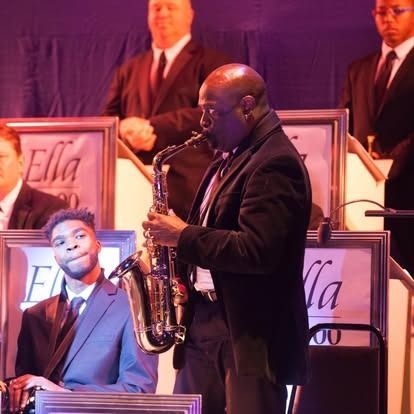 Saxophonist performing on stage with band, wearing black suit, playing saxophone. Other band members in suits are visible.