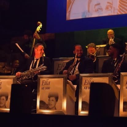 Jazz band performing onstage, in front of framed portraits. Musicians wear suits, some play saxophones and trumpets.