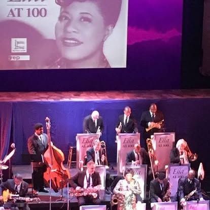 A jazz band performs on stage with a screen showing Ella Fitzgerald. Musicians in suits play instruments.