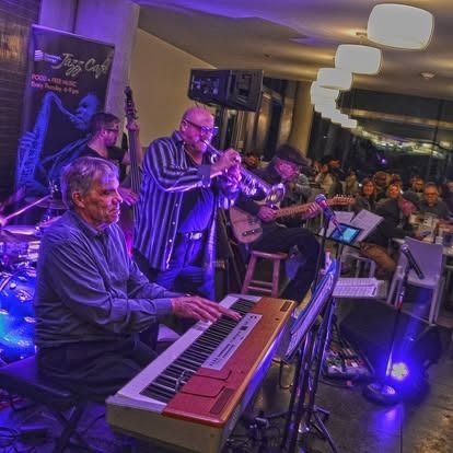Jazz band performing at a cafe: keyboardist, trumpeter, guitarist, bassist, and drummer. Audience in background.