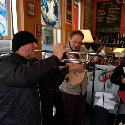 Two men play trumpets indoors. One wears a black cap and coat. The other wears a tan jacket. A bar is in the background.