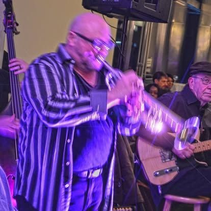 Man playing trumpet in a jazz band, with upright bass and guitar. Purple stage lighting.