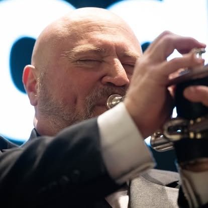 Bald man playing a trumpet, eyes closed in concentration, wearing a black suit.