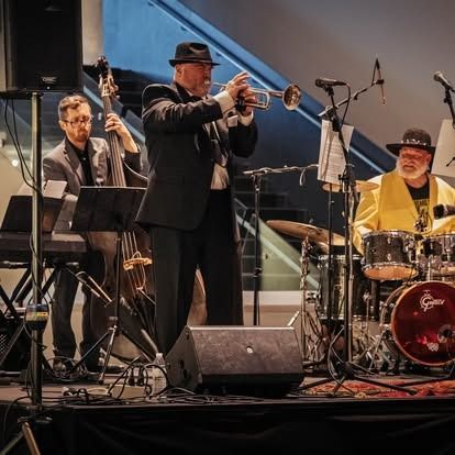 A jazz band performing: trumpet player, bassist, drummer, and keyboardist on stage indoors.