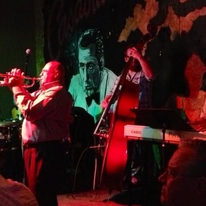 Jazz band performs on stage. Trumpet player, bassist, and keyboardist with an eerie mural backdrop.