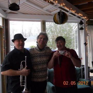 Three men pose inside. One holds a trumpet, another a camera. A relaxed setting.