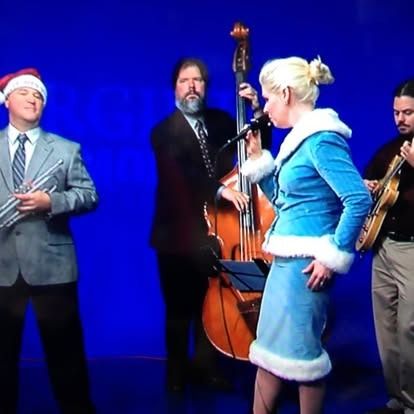 A band of four people performing on a blue background. A woman sings in a light blue outfit. A man plays a standup bass.