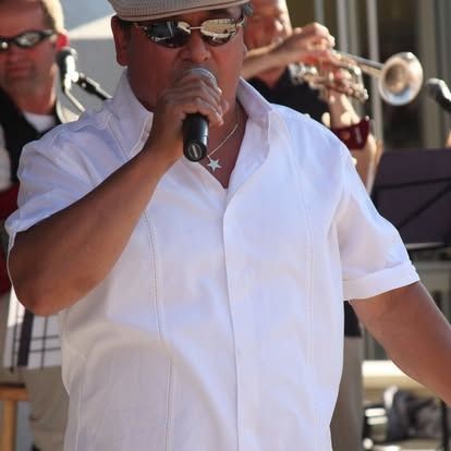 Man singing into a microphone on stage, wearing sunglasses, white shirt, and cap; band in background.