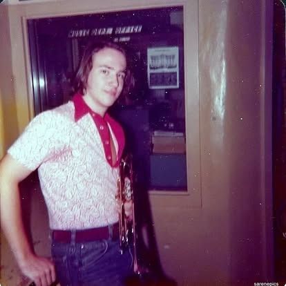 Man in patterned shirt and jeans, holding a trumpet, standing by a door.