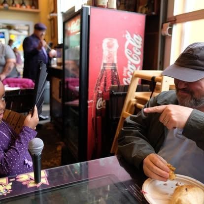 A young girl with a microphone and a man pointing to a plate of food, near a Coca-Cola refrigerator.