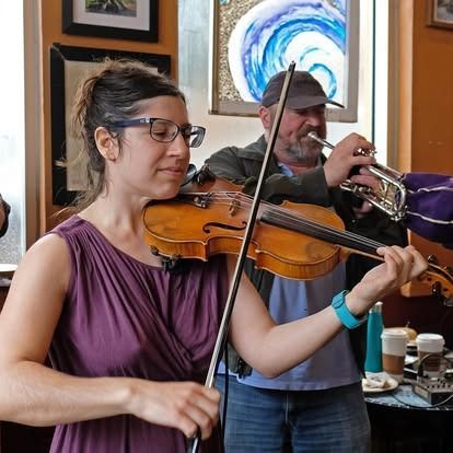 Woman plays violin; man plays trumpet in a cafe. The woman wears glasses and a purple dress.