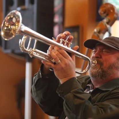 Man playing a silver trumpet, wearing a dark cap and jacket, indoors.