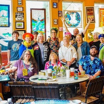 Group of people smiling at a cafe table, colorful clothing, artwork on walls, windows.