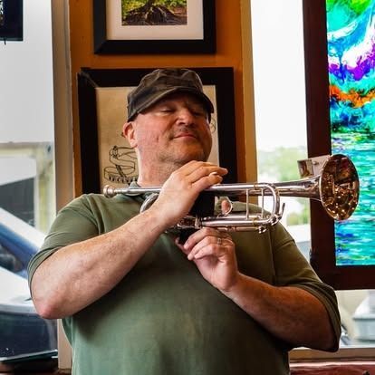 Man in green shirt and cap, holding a silver trumpet, smiling, indoors.